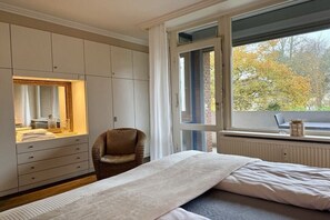 A bedroom with a bed, a chair, a wardrobe, and a window with a view of trees. - Holiday apartment with 1 bedroom (Travemünde)