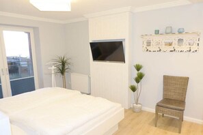 A bedroom with a bed, a television mounted on the wall, a chair, and a shelf with decorative items. - Beach Palace Apt. 108 (Cuxhaven)