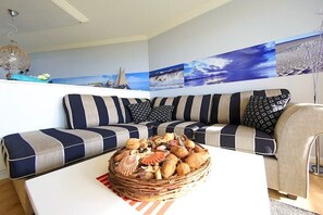 Un salon moderne avec des canapés à rayures, un panier en osier rempli de coquillages et un mur orné d’œuvres d’art sur le thème de la marine.