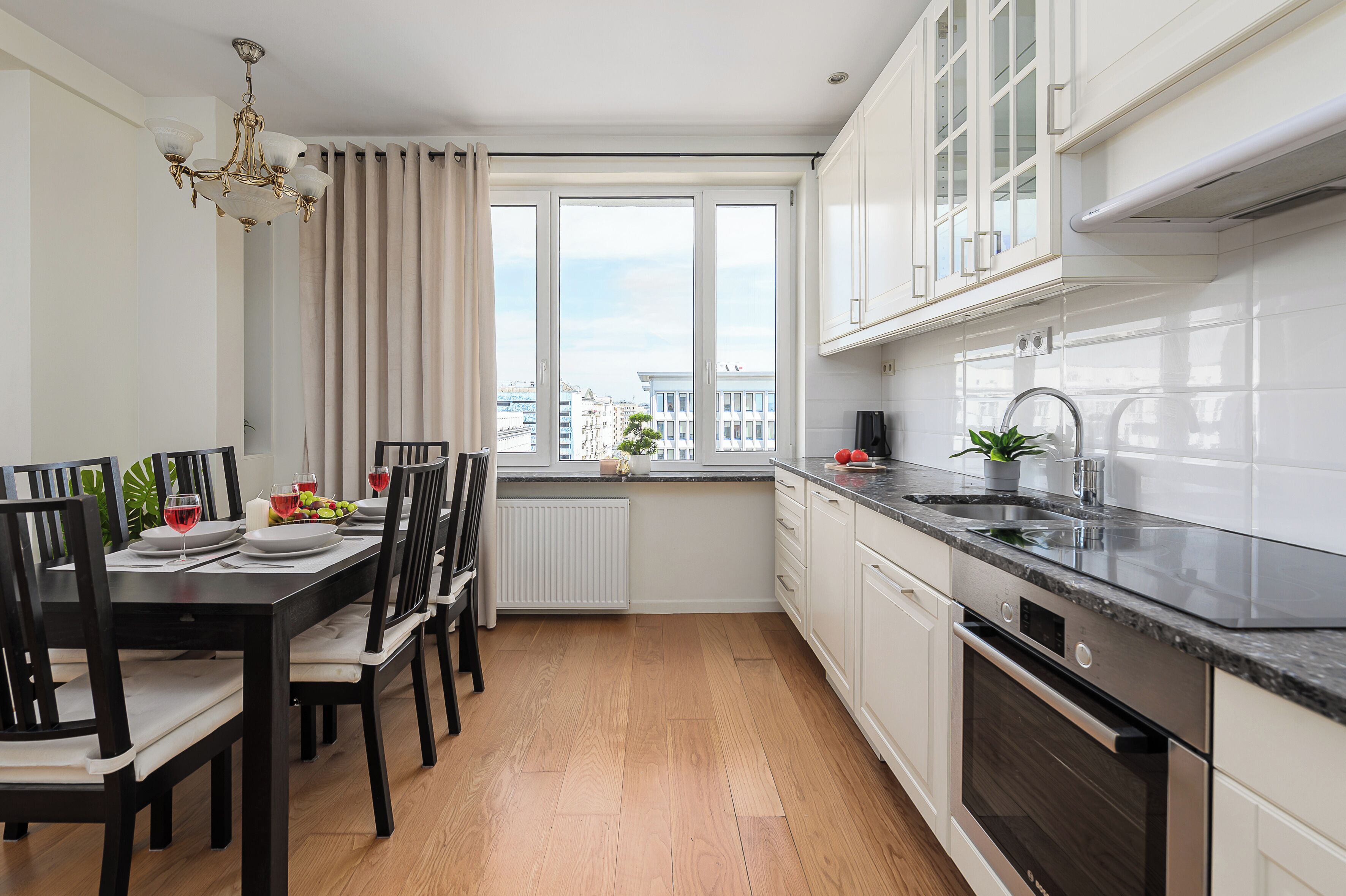 A modern kitchen with a dining area, featuring a large window, white cabinetry, and a black countertop.