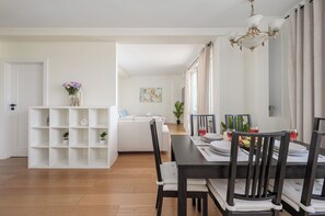Een eethoek met een houten tafel, stoelen en een kroonluchter. Achtergrond is een witte bank en een boekenkast met planten.