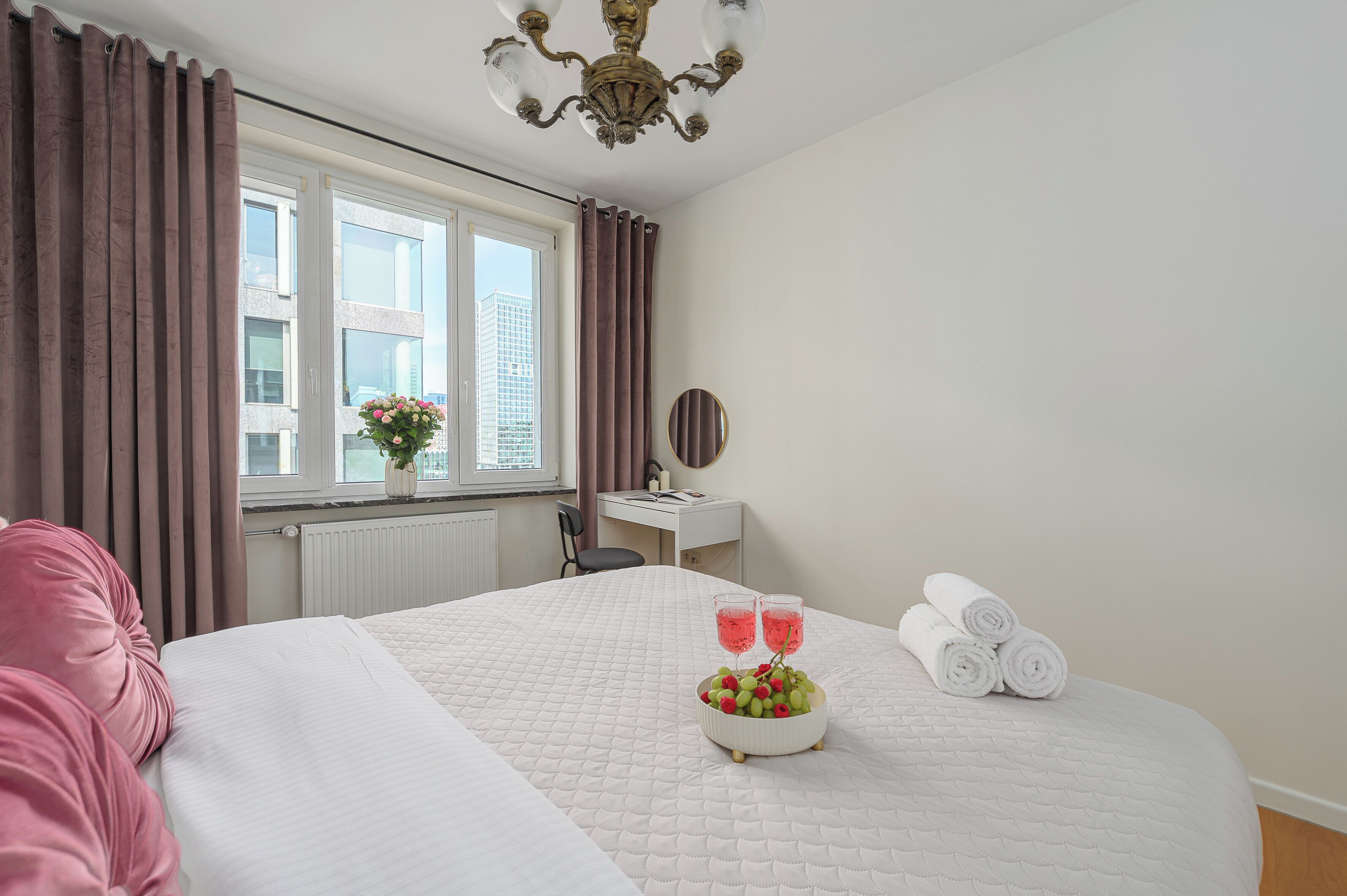 A hotel room with a bed, a desk, a window with curtains, a chandelier, and a bowl of fruit on the bed.