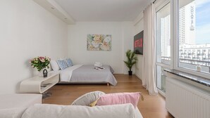Un dormitori modern amb un llit, un sofà, una finestra que dona a una vista de la ciutat i una obra d'art penjada a la paret.