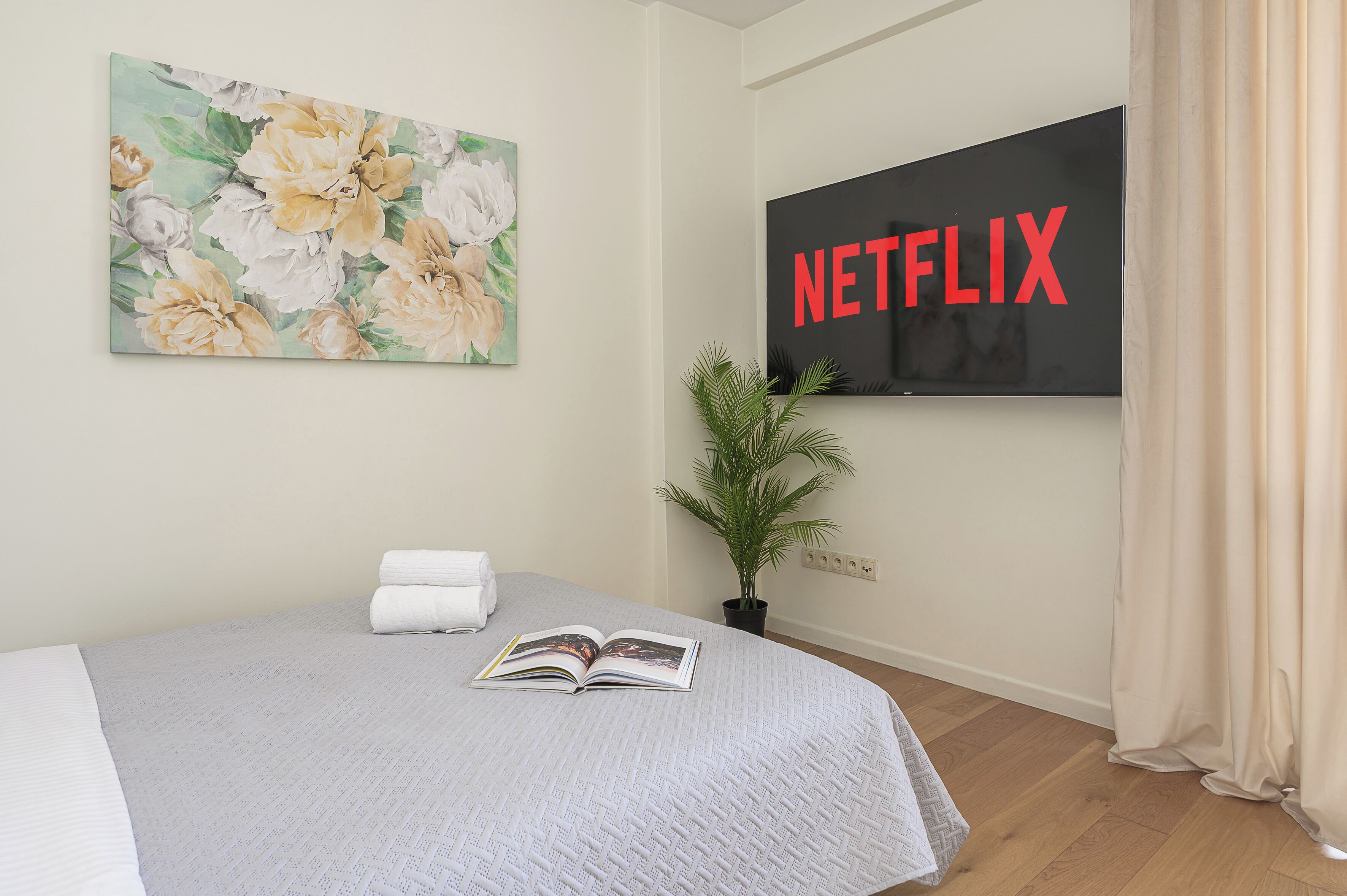 A hotel room with a bed, a painting, a potted plant, a book, and a TV screen displaying the Netflix logo.