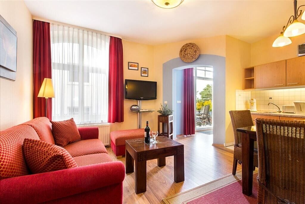 Uma sala de estar com um sofá vermelho, uma mesa de centro, uma TV de tela plana e uma área de cozinha com armários de madeira e um micro-ondas.