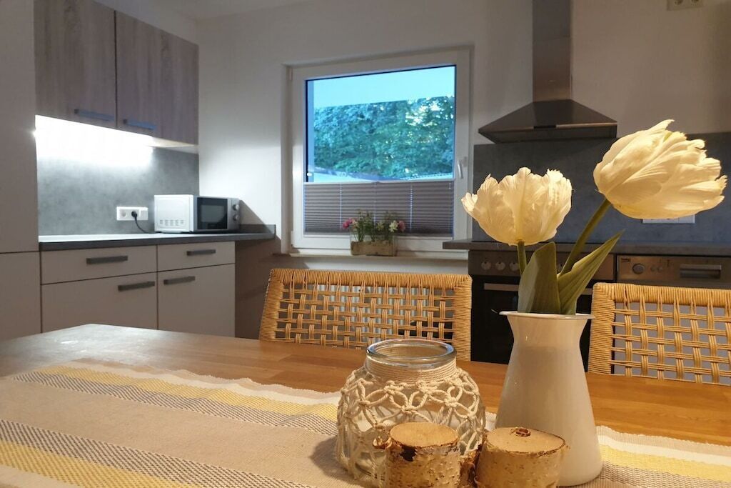 Une cuisine moderne avec une table à manger, des chaises, une fenêtre donnant sur un beau paysage et un vase rempli de fleurs.