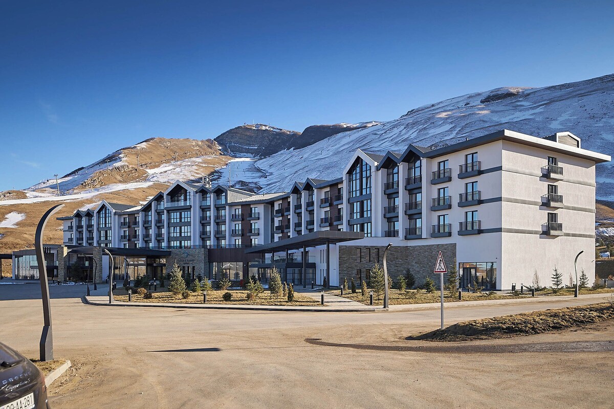 A multi-story hotel with a modern architectural design, featuring balconies and large windows, situated in a mountainous area with snow-covered peaks.