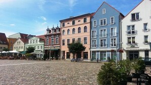 Uma praça de paralelepípedos com edifícios históricos, mesas ao ar livre e vasos de plantas.