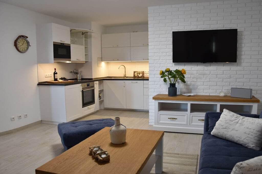 A modern kitchen with white cabinets, a microwave, and a sink. There's a flat-screen TV mounted on a white brick wall, a wooden coffee table with decorative items, and a blue sofa with cushions.