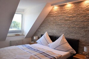 A bedroom with a sloped ceiling, a brick wall, a bed with pillows, a nightstand, and a lamp. - Holiday apartment with 1 bedroom (Ueckermünde)