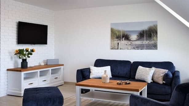 Uma sala de estar moderna com parede de tijolos brancos, uma TV de tela plana montada sobre um console de madeira, um sofá azul escuro com almofadas, uma mesa de centro e uma pintura com tema praiano na parede.