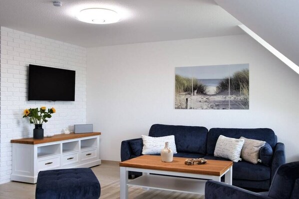A modern living room with a white brick wall, a flat-screen TV mounted above a wooden console, a dark blue sofa with cushions, a coffee table, and a beach-themed painting on the wall. - Holiday apartment with 1 bedroom (Ueckermünde)