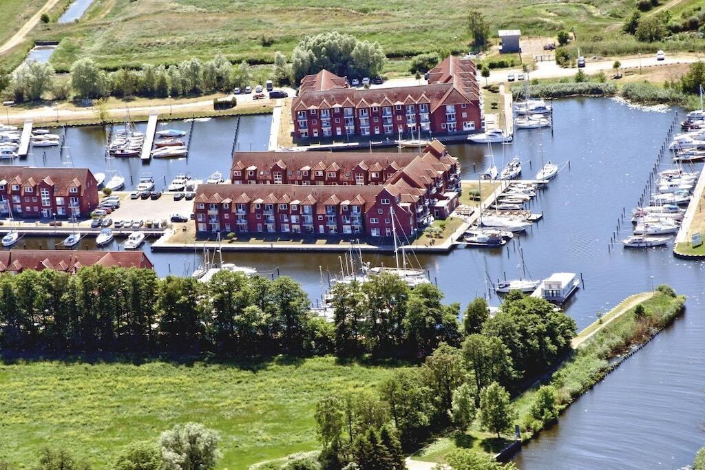 Un port de plaisance où sont amarrés de nombreux bateaux, un grand bâtiment à la façade rouge et une zone herbeuse avec des arbres.