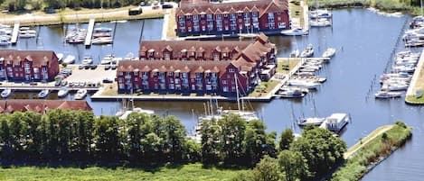 Un port de plaisance où sont amarrés de nombreux bateaux, un grand bâtiment à la façade rouge et une zone herbeuse avec des arbres.