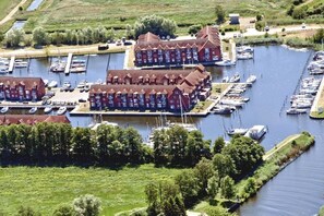 Ein Yachthafen mit mehreren vertäuten Booten, ein großes Gebäude mit roter Fassade und eine begrünte Fläche mit Bäumen.