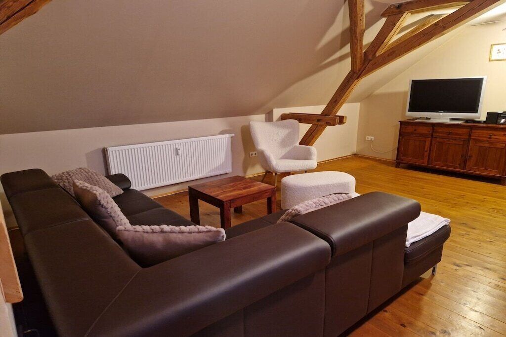 Een woonkamer met een bruine leren bank, een witte fauteuil, een klein houten tafeltje, een witte radiator en een televisie op een houten kast.
