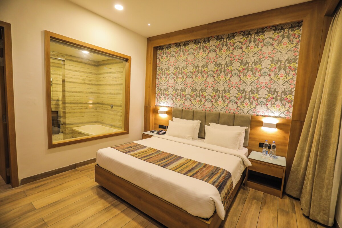 Habitación de hotel con una cama grande, una mesita de noche con lámpara, un espejo y un cabecero estampado con motivos florales.
