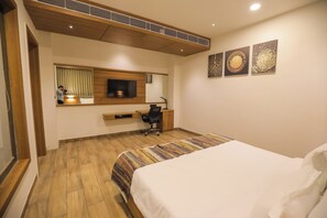 A hotel room with a bed, a desk, a chair, a TV, and artwork on the wall. - RPR RESORT MORBI (Tankara)