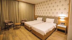 A hotel room with two beds, a small table, two chairs, a nightstand with water bottles, and a curtain.