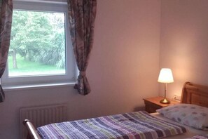 A bedroom with a bed, a nightstand with a lamp, a window with curtains, and a radiator. - Holiday home with 1 bedroom (Westoverledngen)