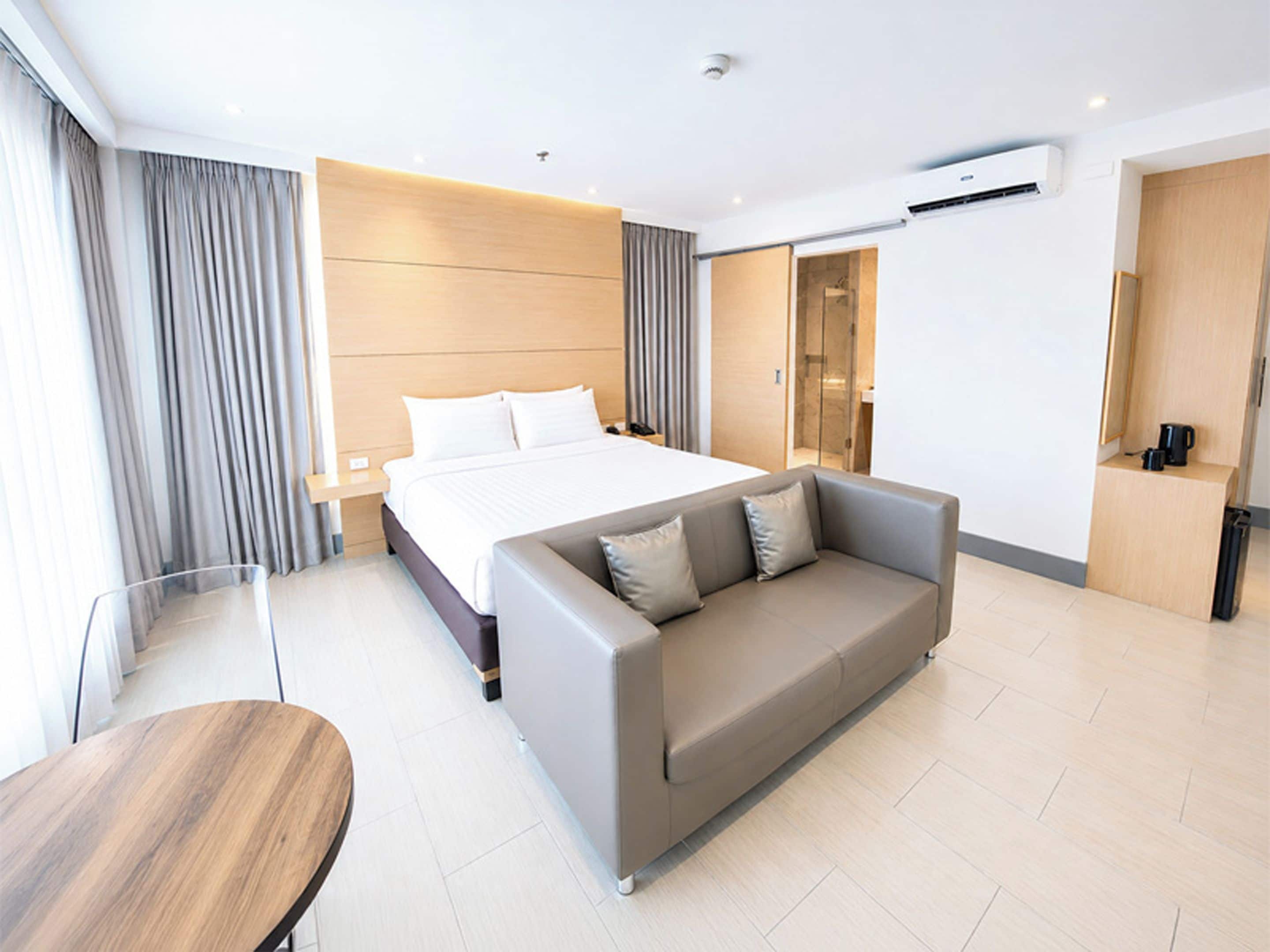 Una habitación de hotel moderna con una cama grande, un sofá, una mesa de madera y un baño visible a través de una puerta de cristal.