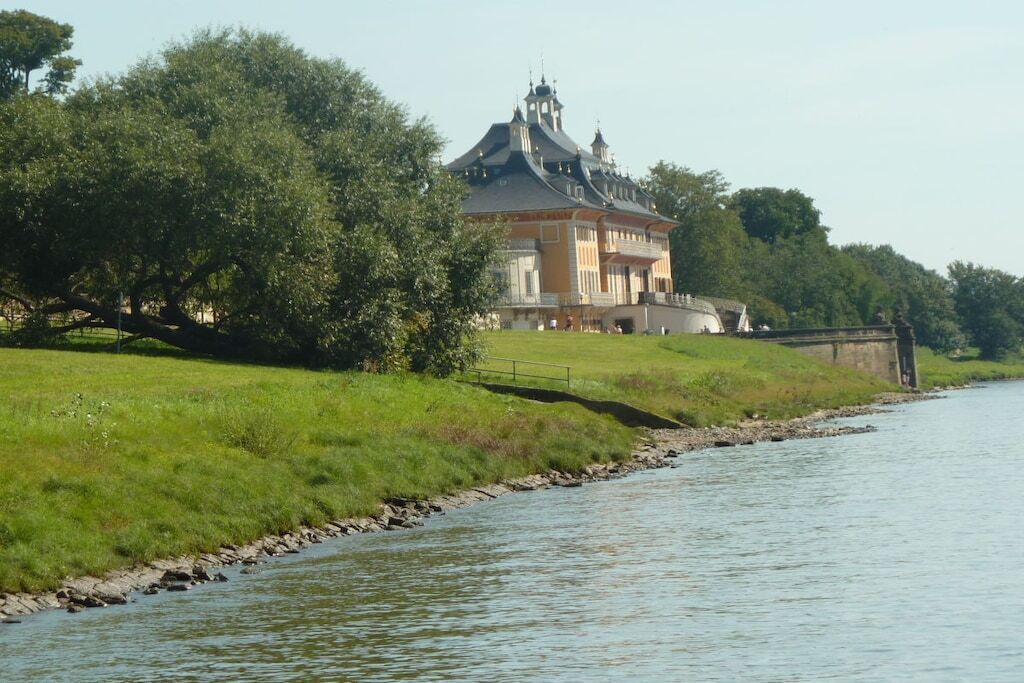 A riverside building with a prominent tower, surrounded by lush greenery and a calm river.
