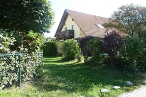 Ein Haus mit einem geneigten Dach, einem eingezäunten Garten und verschiedenen Bäumen und Sträuchern.