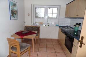 A compact kitchen with a dining table, chairs, a window with curtains, and basic appliances. - Holiday apartment with 1 bedroom (Elbinsel Krautsand)