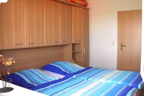 A bedroom with a bed, a bedside table with a lamp, wooden wardrobe, and a door. - Holiday apartment with 1 bedroom (Elbinsel Krautsand)
