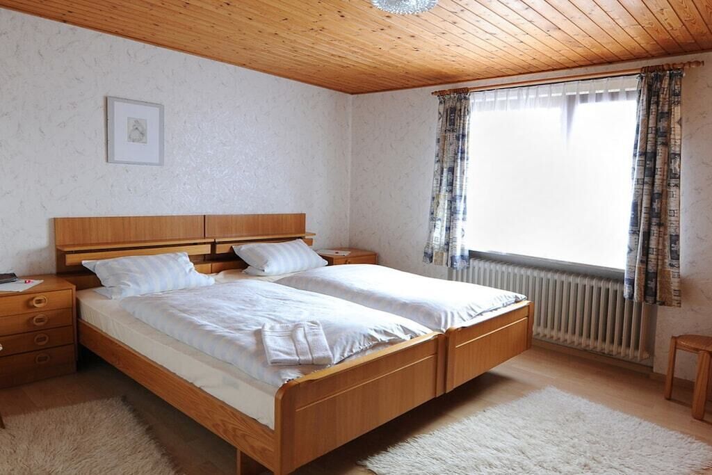 Een slaapkamer met een houten bed, nachtkastje, dressoir, raam met gordijnen en een radiator.