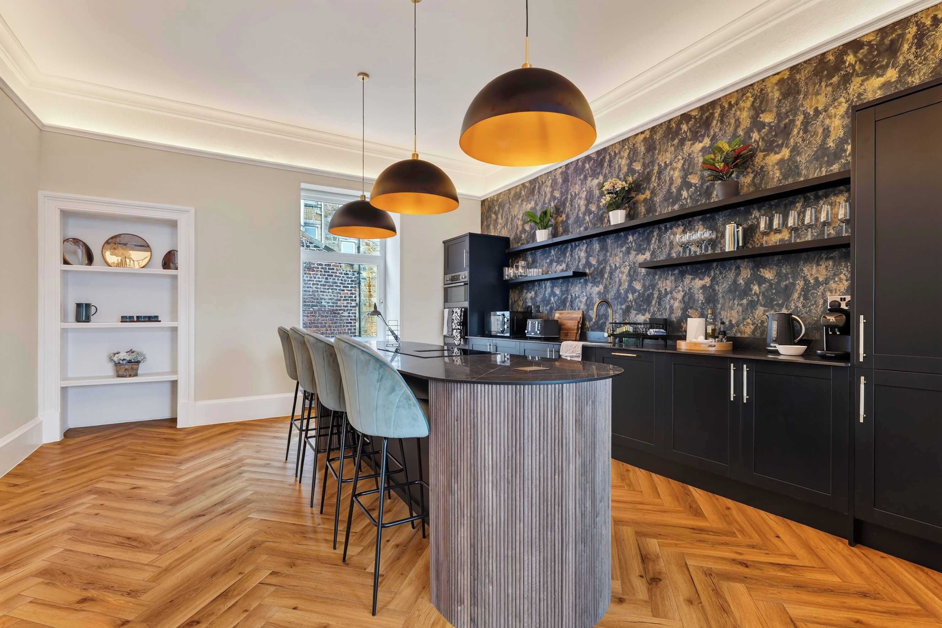 A modern kitchen with a herringbone wood floor, a central island with bar stools, pendant lighting, and a brick wall backsplash.
