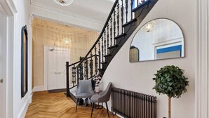 Uma escadaria com balaústres pretos, uma porta branca e um espaço de estar com duas cadeiras e uma mesa.