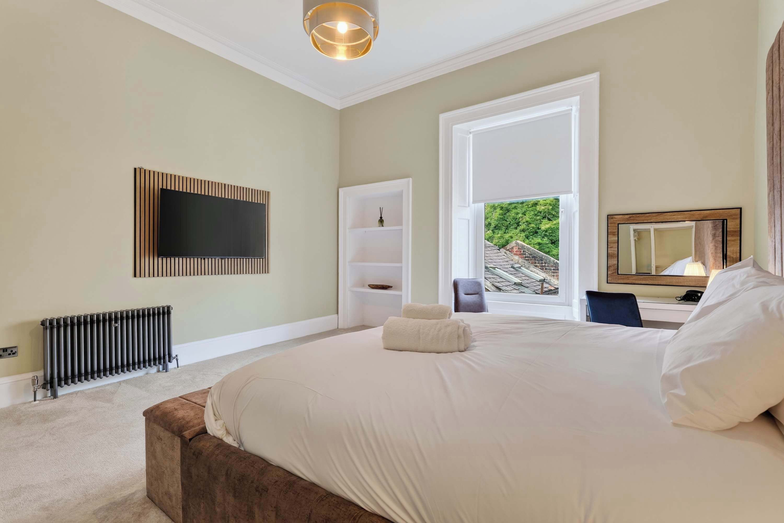 Een slaapkamer met een groot bed, een aan de muur bevestigde televisie, een radiator en een raam met uitzicht op groen.