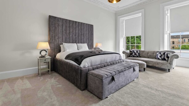 Quarto com uma cama grande, um sofá e um banco. Janela com vista para edifícios e árvores.
