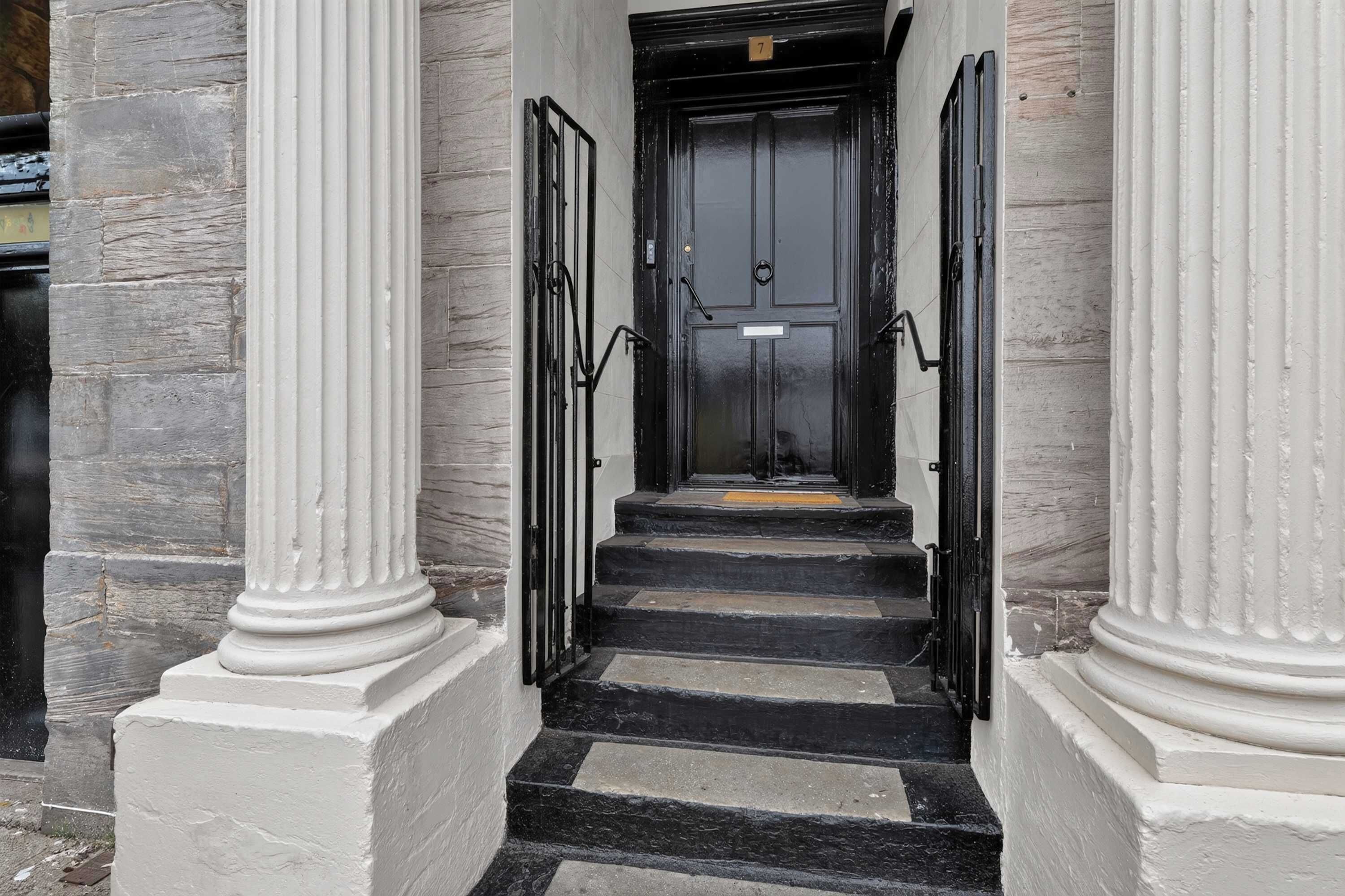 A staircase leading to a black door with a number plate, flanked by white columns and a stone wall.