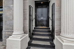 Una escalera conduce a una puerta negra con una placa de número, flanqueada por columnas blancas y un muro de piedra.