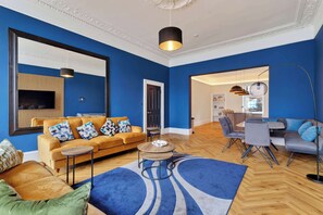 A modern living room with a blue wall, a sofa, a coffee table, and a dining area with a table and chairs. - Spacious 5-Bedroom Glasgow Home Perfect for City Breaks (Glasgow City)