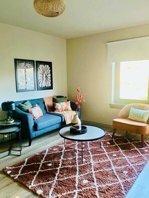 A living room with a blue sofa, a round coffee table, a small side table, a decorative vase with flowers, a brown rug, and a window with a view. - Bright and Central Paisley Flat Fast Airport and Glasgow Links (Renfrewshire)