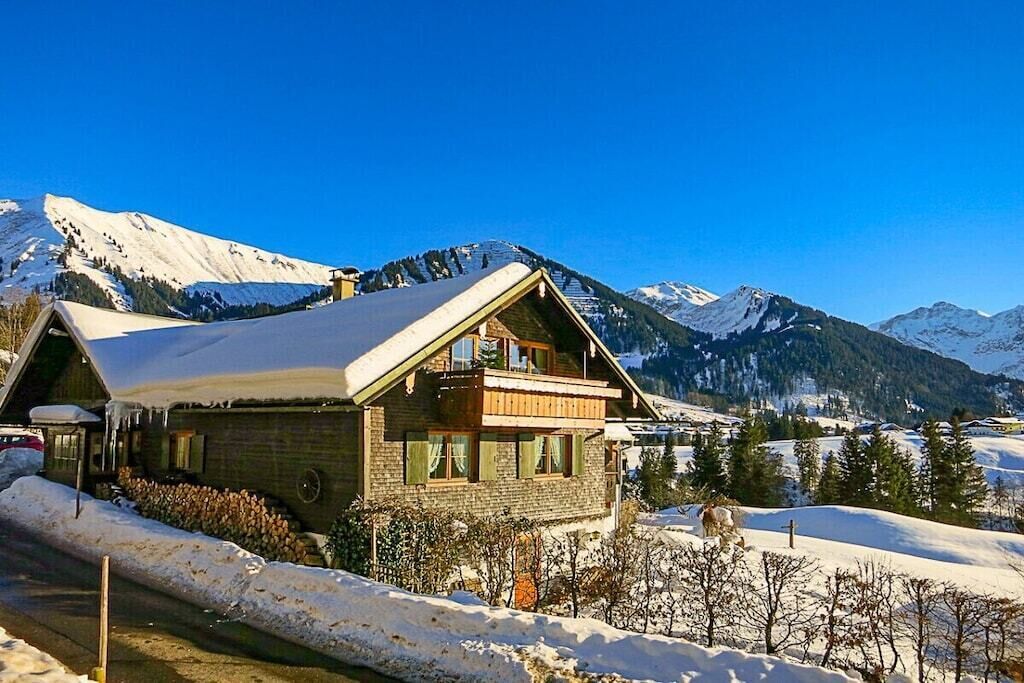Ein Chalet mit geneigtem Dach, Holzfassade und einem Balkon, umgeben von schneebedeckten Bergen.