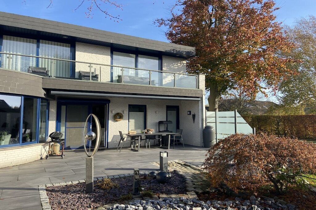 Un bâtiment moderne à deux étages, doté d’un balcon, d’un espace extérieur aménagé pour s’asseoir et d’un jardin agrémenté d’une sculpture métallique décorative.