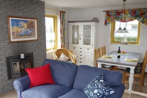 A living room with a blue sofa, a fireplace, a dining table set for two, and a cabinet with glass doors. - Holiday home with 2 bedrooms (Julianadorp aan Zee)