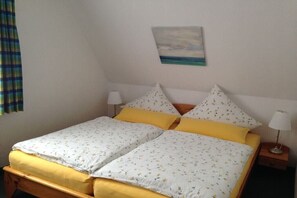 Een eenpersoonsbed met een wit en geel bloemenbeddengoed, twee kussens, een nachtkastje met een lamp, en een schilderij van een strandzicht aan de muur.