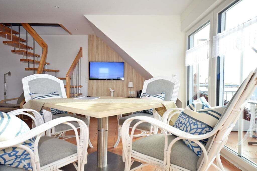 A dining area with wicker chairs and a wooden table, a TV mounted on the wall, and a staircase in the background.