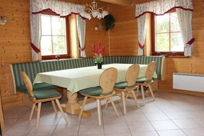 Interior de una cabaña de madera con una mesa de comedor, cuatro sillas y dos ventanas con cortinas.