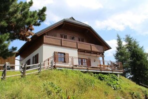 Eine zweigeschossige Holzchalet mit einem Balkon im oberen Stockwerk, umgeben von Bäumen und einer grünen Böschung.