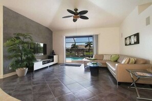 A modern living room with a flat-screen TV, a ceiling fan, a large potted plant, a beige sofa, and a glass coffee table. - Villa in Cape Coral with swimming pool (Cape Coral)