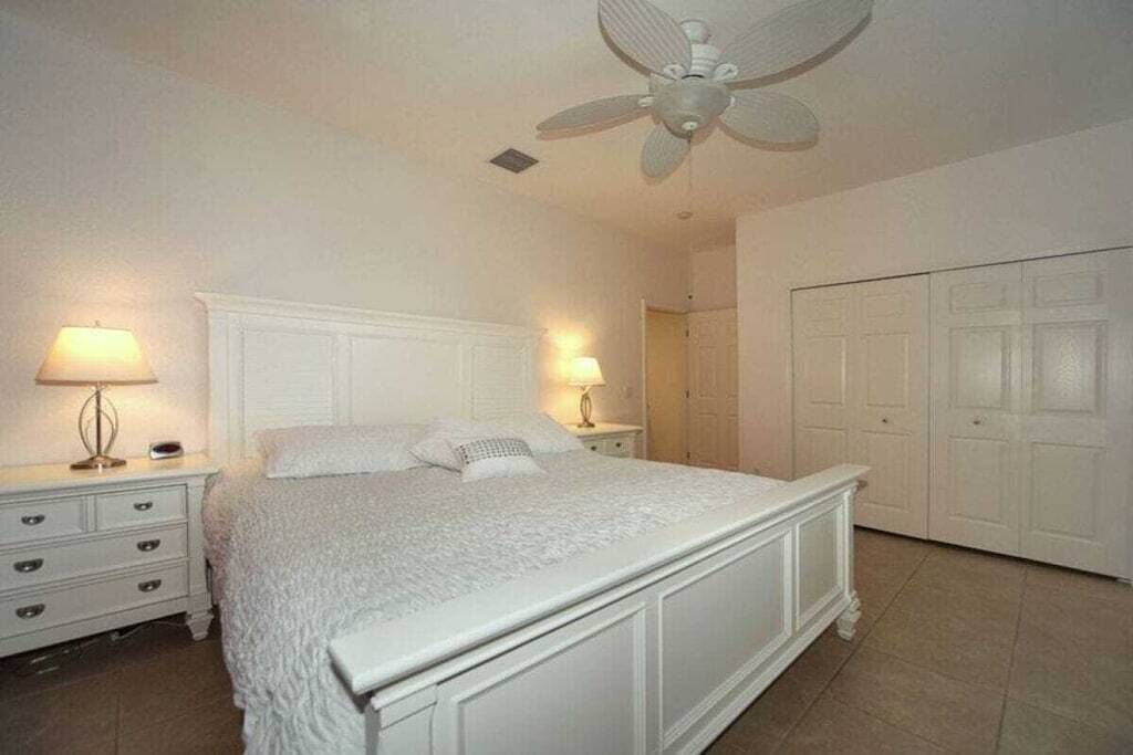 A bedroom with a large bed, two bedside tables with lamps, a ceiling fan, and a white wardrobe.