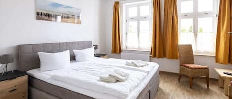Una habitación de hotel con cama, una silla, mesita de noche, lámpara y ventana con cortinas.
