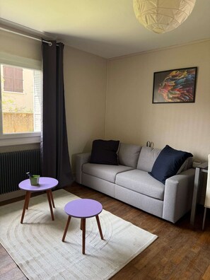A living room with a grey sofa, two round tables, a radiator, a window with curtains, and a wall-mounted painting. - Cozy studio in the heart of Charnay (Charnay-lès-Mâcon)