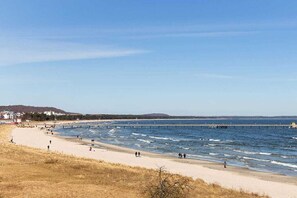 Ein Strand mit einem Pier, der ins Wasser ragt, sandigem Ufer und Gebäuden im Hintergrund.